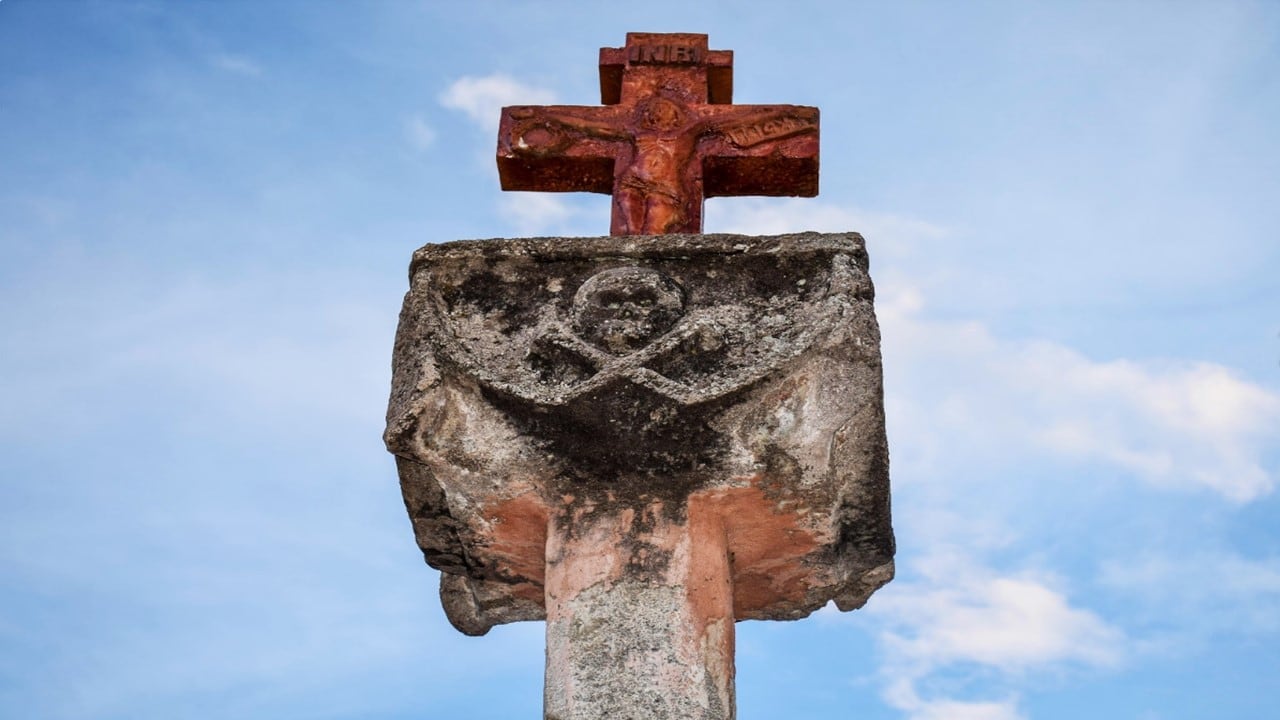 Una cruz de piedra desgastada, que presenta un cristo crucificado y un relieve de calavera con tibias cruzadas sobre un pedestal, se alza contra un cielo azul con nubes blancas.