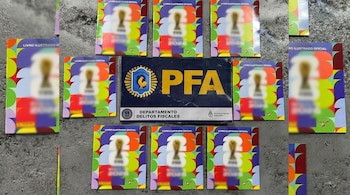 Vista aérea de varios álbumes coleccionables del Mundial y una placa azul de la PFA con 'DEPARTAMENTO DELITOS FISCALES' sobre una superficie gris.