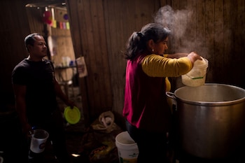 Una mujer vierte arroz de un cubo a una olla humeante, mientras un hombre sostiene un plato y un vaso en una cocina con paredes de madera