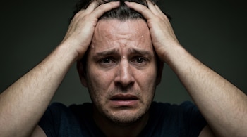 Primer plano de un hombre con el pelo oscuro y sudor en la frente, sujetándose la cabeza con ambas manos, con expresión de miedo. Fondo oscuro.