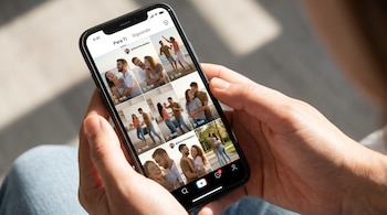 Primer plano de dos manos sosteniendo un smartphone negro. La pantalla muestra una interfaz de red social con una cuadrícula de videos de parejas felices en diversas situaciones.