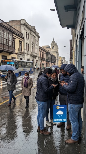 Personas abrigadas y con mascarillas caminan por calles mojadas de Lima; un grupo consulta móviles y se ve un pronóstico de Senamhi de 13°C.