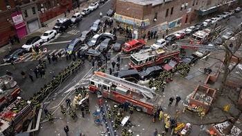 Vista aérea de calle urbana con camiones de bomberos, bomberos, personal de emergencia y vehículos estacionados. Edificios y personas en la escena del incidente