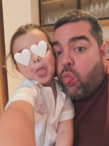 Hombre barbudo y niña con los ojos tapados por corazones blancos sacan la lengua, en un plano cercano de sus rostros