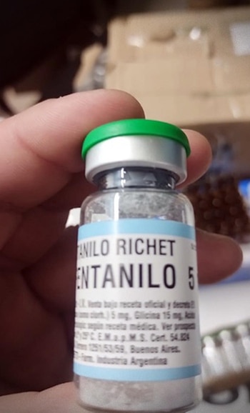 Mano sosteniendo un pequeño vial de vidrio transparente con una tapa verde y plateada. La etiqueta blanca dice 'FENTANILO RICHET' y 'FENTANILO 5'