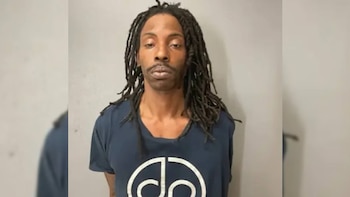 Hombre con cabello trenzado tipo rastas, bigote y barba corta, camiseta azul con un logo circular blanco, de pie frente a una pared clara