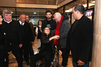 Charly García en silla de ruedas sonríe, usando gafas y chaqueta negra, interactuando con Pedro Aznar, quien lleva una bufanda roja, en un vestíbulo