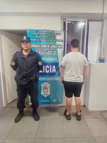 Oficial de policía uniformado de pie junto a un hombre de espaldas, con camiseta blanca y pantalón corto, frente a un cartel de la Policía de Buenos Aires