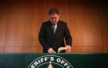 Hombre de traje oscuro y corbata clara, con gafas y bigote, de pie frente a una mesa con un mantel verde con el logotipo "Sheriff's Office", revisa papeles
