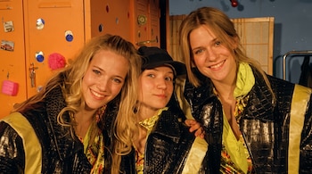 Tres mujeres con chaquetas negras de textura de cuero y detalles amarillos posan cerca de un casillero naranja.