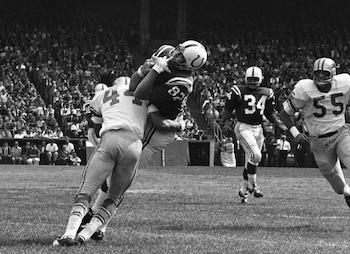 Fotografía en blanco y negro de un partido de fútbol americano. Un jugador con el dorsal 82 (uniforme oscuro) atrapa el balón mientras es tackleado. Otros jugadores y espectadores en las gradas son visibles