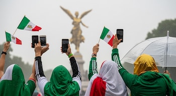Personas con camisetas verdes y ropa tricolor. Sostienen celulares y banderas de México. Lluvia y paraguas. Ángel de la Independencia desenfocado.