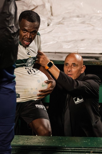 Hombre de piel oscura con camiseta de Coritiba, sujetado por un hombre calvo de traje oscuro, junto a una barandilla