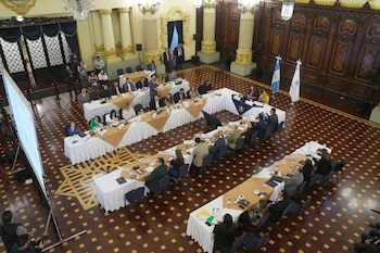 Personas sentadas en mesas dispuestas en rectángulo dentro de un salón con columnas, lámparas y una bandera de Guatemala