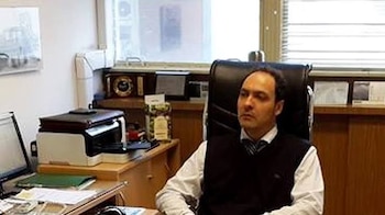 Un hombre con camisa y chaleco sentado en una silla de cuero negro junto a un escritorio con monitor e impresora