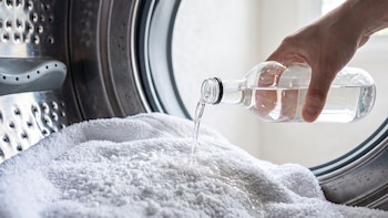 Mano de persona vierte líquido transparente de botella de vidrio sobre toallas blancas dentro del tambor de lavadora