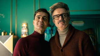 Tristán Ulloa, con jersey burdeos, y Pedro Alonso, con cárdigan marrón y gafas, posan sonrientes frente a una pared verde esmeralda con decoración dorada