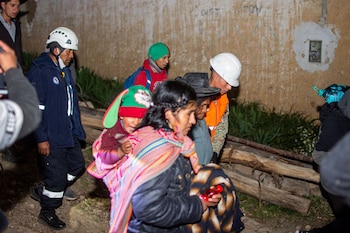Grupo de personas: personal de rescate con cascos y chalecos, civiles con niños y vestimenta tradicional. Caminan junto a una pared dañada y terreno irregular