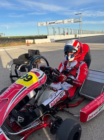 Un piloto con casco y traje de competición está sentado en un kart rojo y blanco junto a una silla de ruedas en una pista de carreras