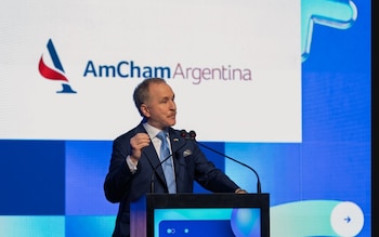 Un hombre con traje azul y corbata habla tras un podio con micrófonos, frente a una pantalla con el logotipo de AmCham Argentina
