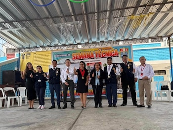 Nueve personas posan en un escenario escolar con el pulgar levantado delante de un cartel de la Semana Técnica del colegio Kumamoto