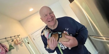 Un hombre maduro y sonriente sostiene con ambas manos a un perro pequeño equipado con un arnés dentro de una cocina