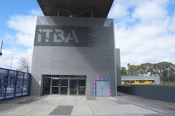 Edificio gris con la sigla ITBA en la fachada, puertas de vidrio en la entrada principal y una reja azul en el sector izquierdo