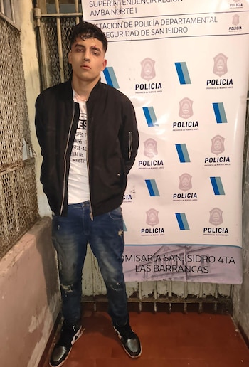 Un hombre joven con campera negra y pantalón de jean posa frente a un banner oficial de la Policía de la Provincia de Buenos Aires