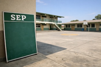 Pizarrón verde con logo SEP apoyado en pared, frente a patio de escuela pública mexicana vacía con edificios de dos pisos y aro de baloncesto bajo cielo azul.