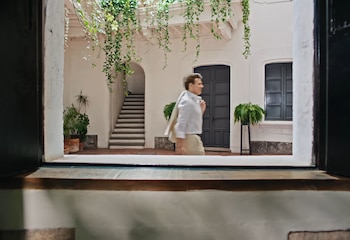 Un hombre vestido con camisa blanca camina por un patio de paredes blancas con plantas colgantes, escaleras y puertas oscuras