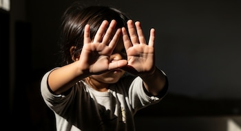 Primer plano de una niña con cabello oscuro. La niña tiene las manos abiertas cubriendo su rostro, que está en sombra. Viste una camiseta gris. Fondo oscuro.