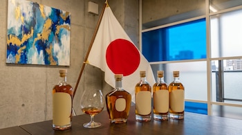 Varias botellas de ron y una copa con ron sobre una mesa de madera oscura. De fondo, una bandera de Japón en un asta y una pintura abstracta azul y dorada.