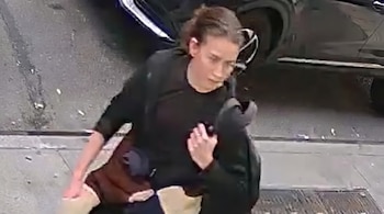 Persona con ropa oscura y el cabello recogido que transita por una acera junto a un automóvil negro.