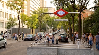 Cartel de la estación de metro Guzmán el Bueno en una calle con árboles, vehículos estacionados y personas caminando.