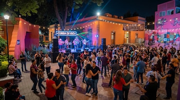 Varias parejas bailan en una plaza iluminada de noche frente a un escenario con músicos y un letrero que dice Cumbia en Vivo.