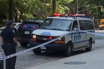 Una camioneta y un SUV de la policía de Nueva York con luces de emergencia encendidas y cinta policial en una calle, con un oficial de espaldas y árboles al fondo