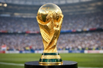 Primer plano de la Copa Mundial de la FIFA, dorada y brillante, sobre un pedestal oscuro. En el fondo borroso, se ven un estadio de fútbol y espectadores.