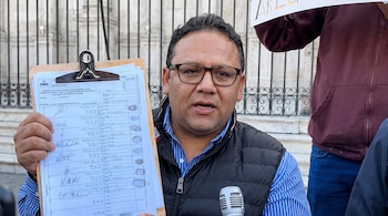 Hombre con gafas y chaleco oscuro sostiene un portapapeles con documento de ONPE y firmas. Micrófono gris y reja metálica de fondo.