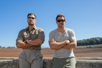 Dos hombres con gafas de sol y brazos cruzados posan al aire libre, con un cielo azul brillante, un campo de tierra y árboles al fondo