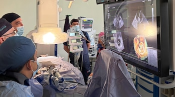 Cinco profesionales médicos con cubrebocas y gorros en un quirófano, con un paciente, equipos de monitoreo y una pantalla con imágenes médicas.