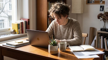 Un joven de cabello desordenado, con suéter, sentado en un escritorio de madera trabajando en una laptop plateada con una mini planta al lado.