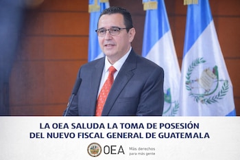 Hombre de traje oscuro, corbata roja y gafas hablando en un podio con micrófonos, flanqueado por dos banderas de Guatemala al fondo.