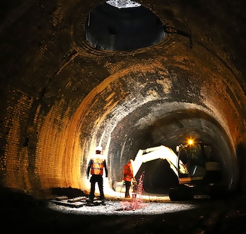 Dos trabajadores con cascos y chalecos de seguridad, una excavadora y luces operan dentro de un túnel de ladrillo abovedado con una abertura superior