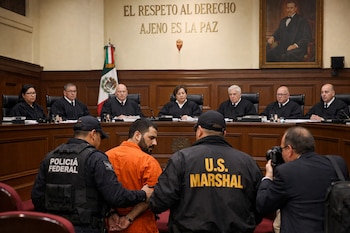 Un hombre esposado con traje naranja es escoltado por dos oficiales, uno de la Policía Federal y otro de U.S. Marshal, frente a siete jueces con toga en un tribunal.