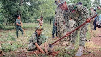 Una mujer uniformada coloca una planta en la tierra mientras un hombre sostiene una azada en un entorno forestal