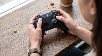 Manos de una mujer con anillo y reloj, sosteniendo un control de videojuegos Xbox en una mesa de madera con migas y un vaso de café.