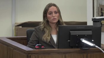 Una mujer con traje oscuro permanece sentada en el estrado de un tribunal frente a un monitor de computadora