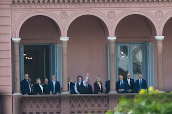 Varias personas, incluyendo hombres y mujeres, de pie en el balcón de un edificio rosado con ventanas arqueadas; uno de ellos saluda con la mano