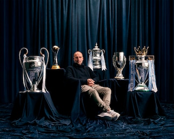 Pep Guardiola, un hombre calvo con suéter de cuello alto oscuro y pantalones claros, está sentado entre cinco trofeos de fútbol, sobre un fondo de cortinas oscuras