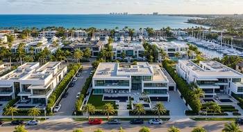 Vista aérea de un barrio costero de lujo con mansiones modernas, piscinas, palmeras, autos en la calle y yates en una marina, frente al océano.